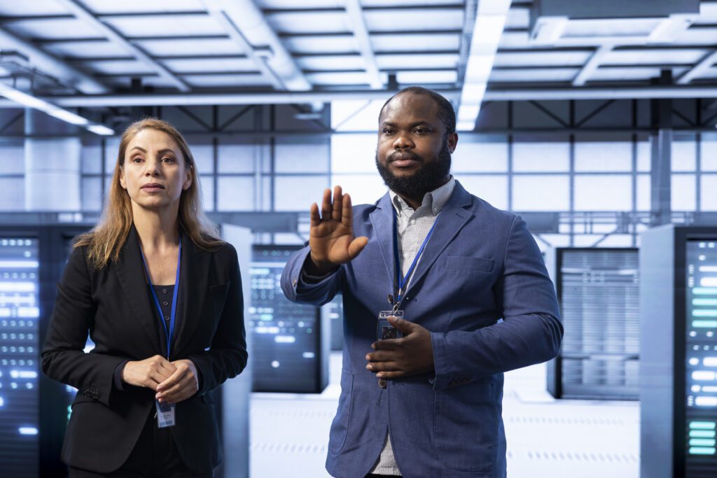 Two professionals in a server room, one gesturing a stop sign to emphasize data protection. They both wear ID badges and formal attire.