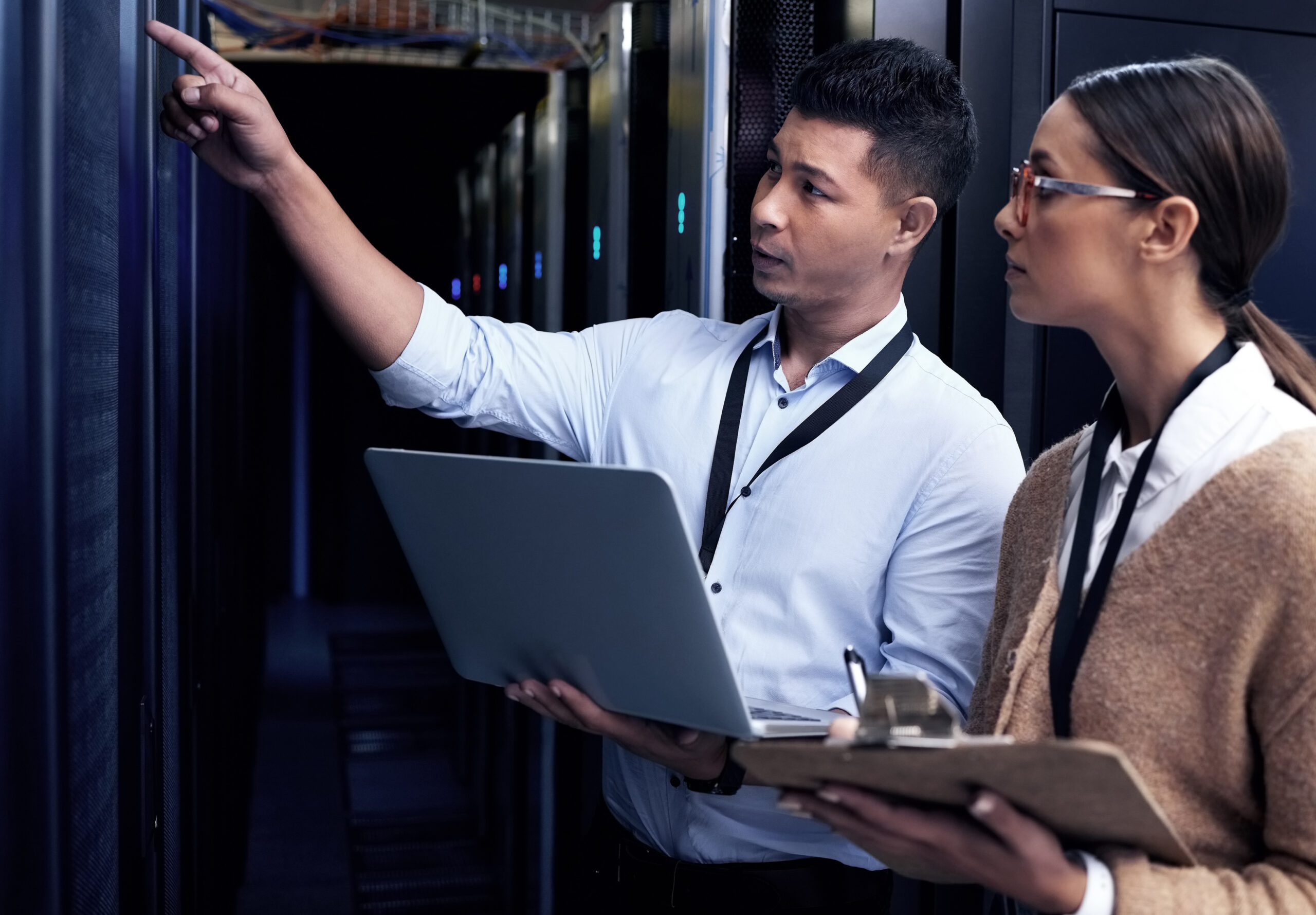 Two people in a server room: one points at equipment while the other holds a clipboard, discussing IT infrastructure. A laptop is visible, likely accessing cloud computing systems.