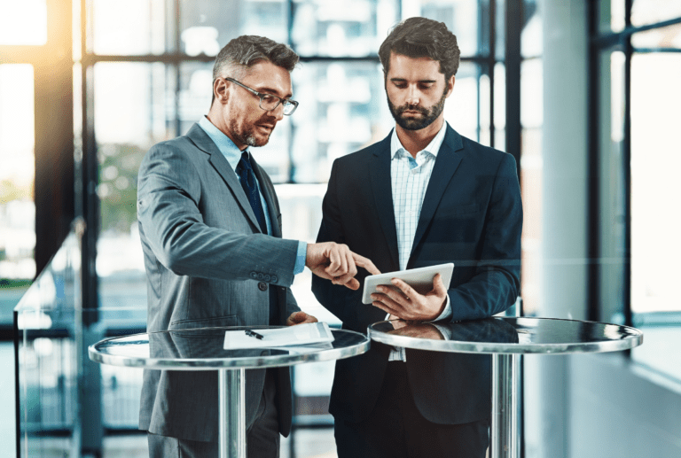 Two men in business attire stand at a round glass table, discussing data security on a tablet in a modern office setting.