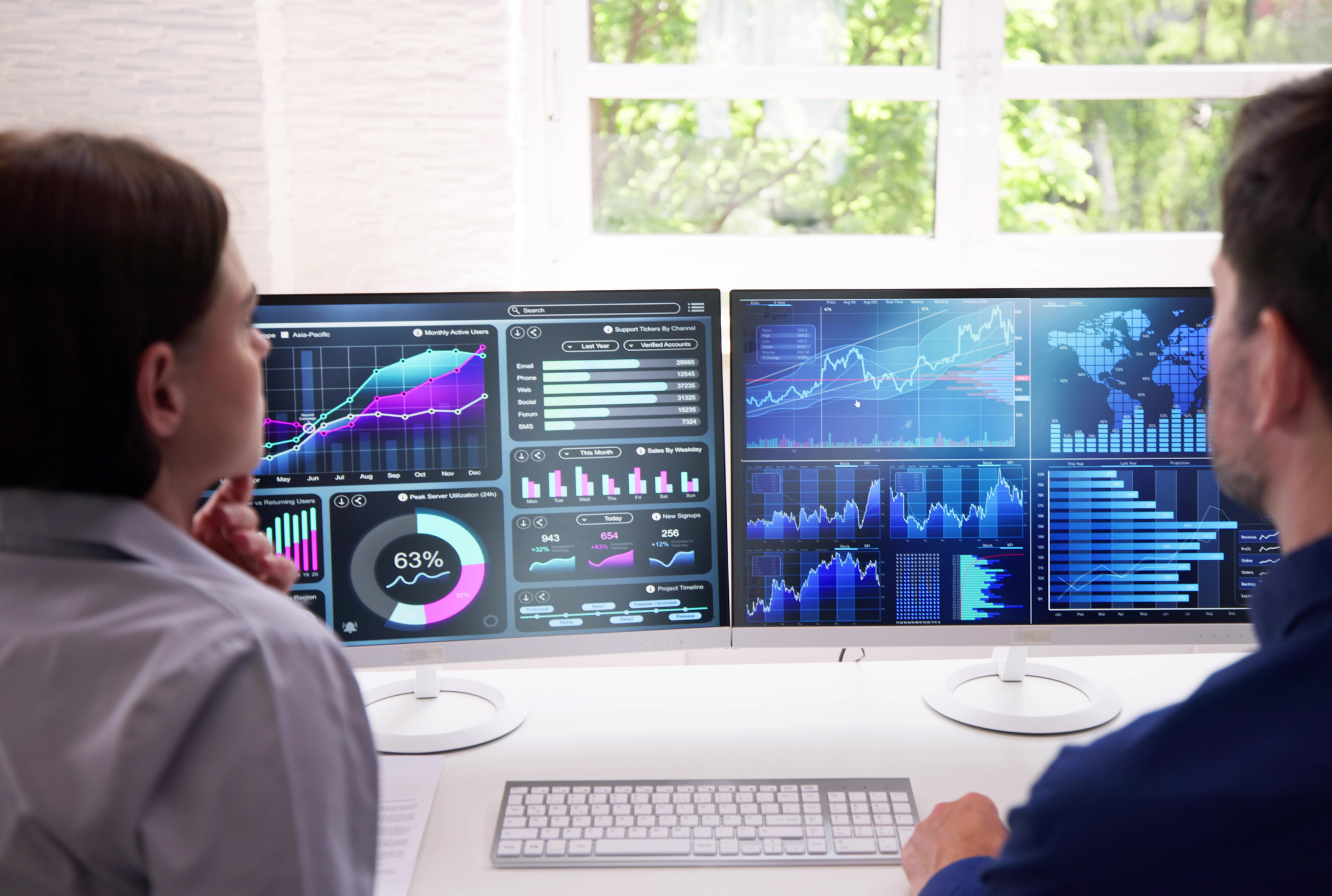 In a bright office setting, two people analyze data on dual monitors displaying charts, graphs, and maps. The green glow from the screens casts an energizing aura as they delve deeper into their analysis.