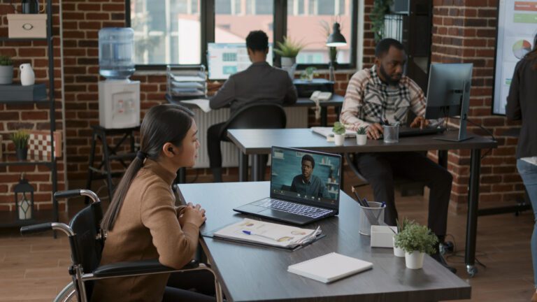 People working in an office with brick walls, using computers and video conferencing. A woman is focused on a virtual meeting on her laptop, seamlessly utilizing remote access for efficiency.