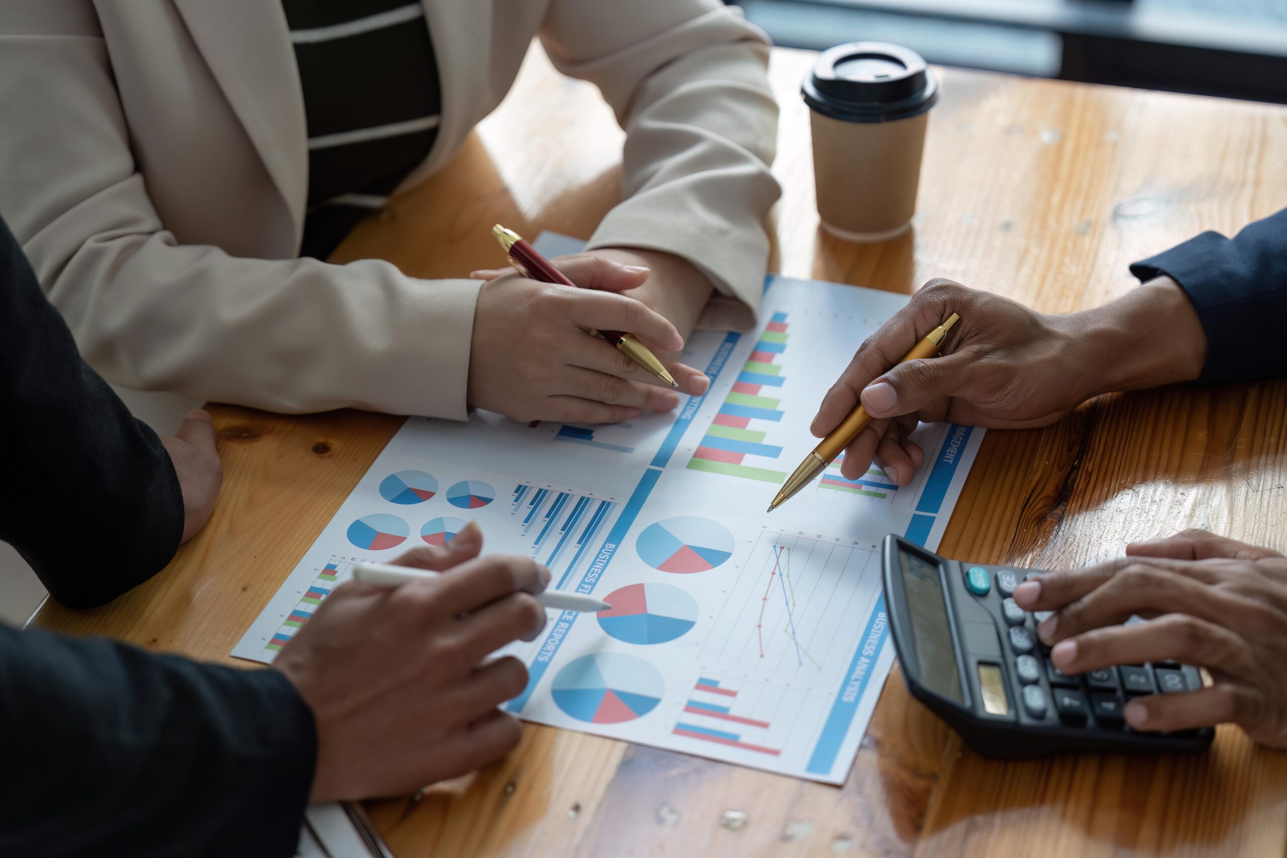 Three people discuss charts and graphs on a wooden table, hands holding pens, a calculator, and a coffee cup. The conversation hints at digital marketing strategies as vital tools for business transformation.