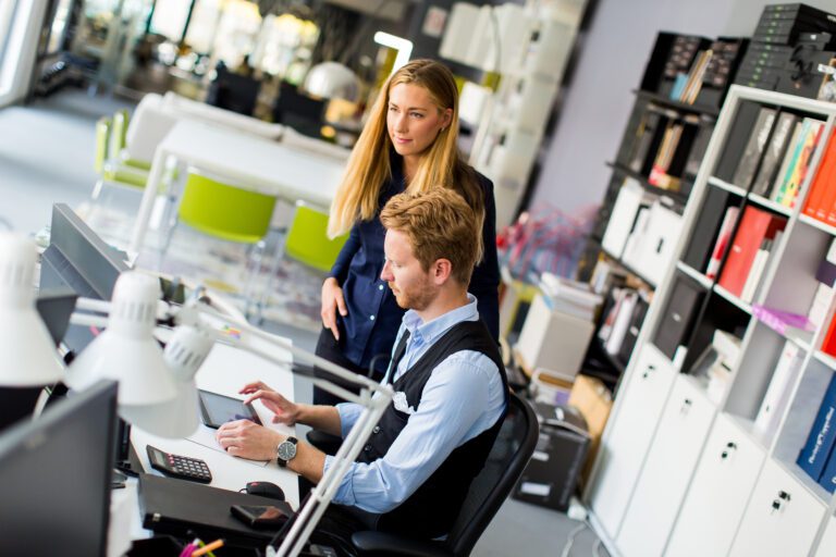 Two people collaborate in a modern office. A seated person uses software on the computer while another stands nearby, discussing or observing. Shelves and office supplies are visible in the background, showcasing an efficient workspace with all the necessary tools for success.