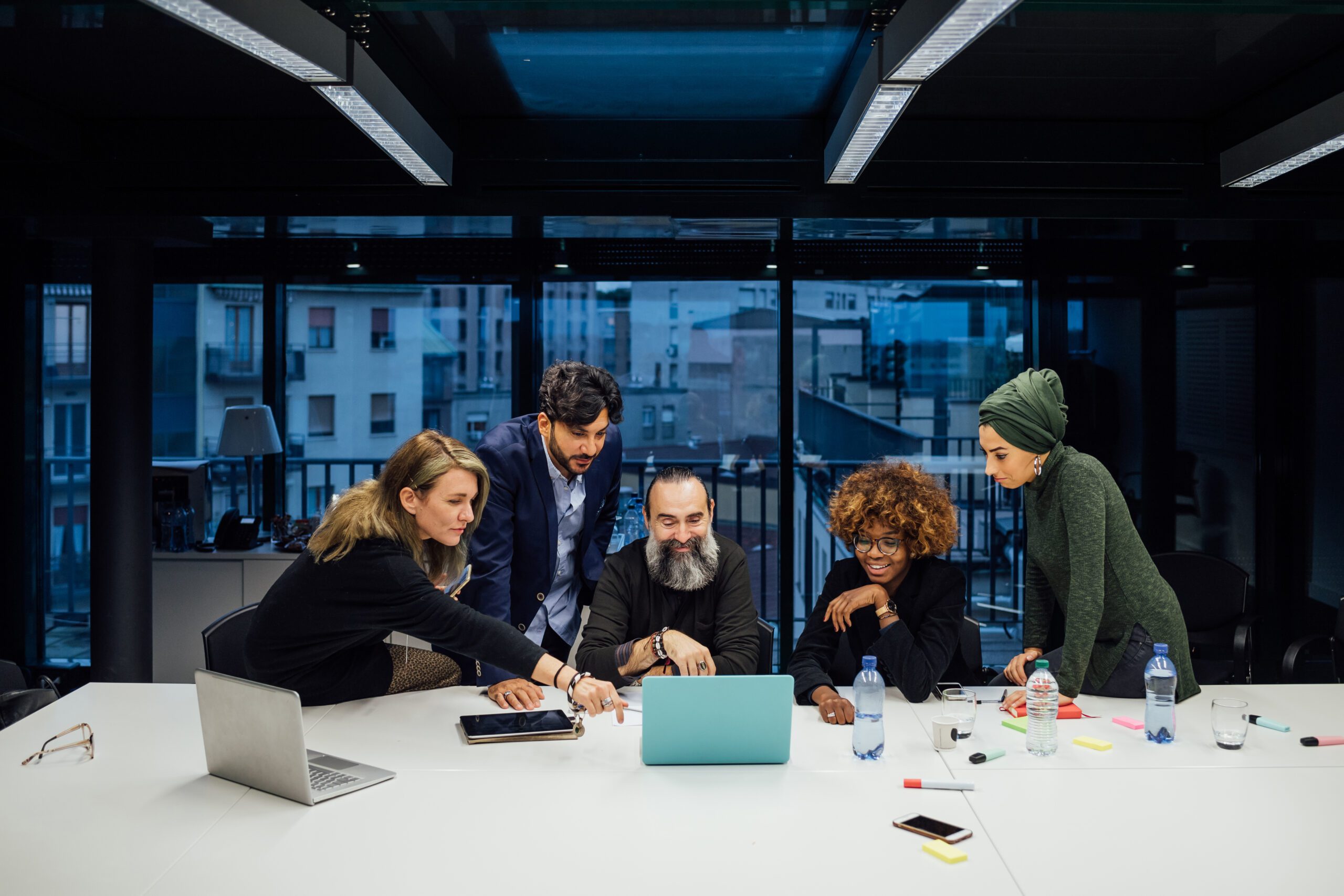 Five people collaborate around a table with laptops and papers in a modern office setting, utilizing cutting-edge software tools to enhance their teamwork.