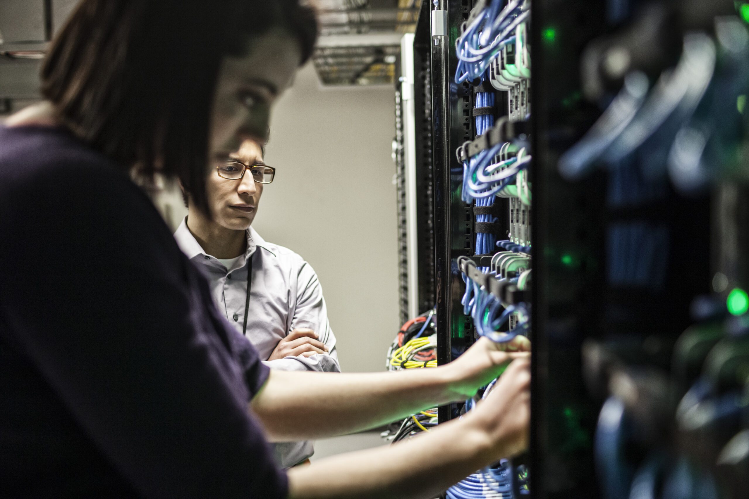 Two people in a server room, examining cables and network equipment, ensure seamless connectivity as part of their managed IT services.