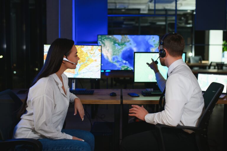 Two people in a control room, focused and wearing headsets, expertly manage the risk as they analyze computer screens displaying detailed maps.