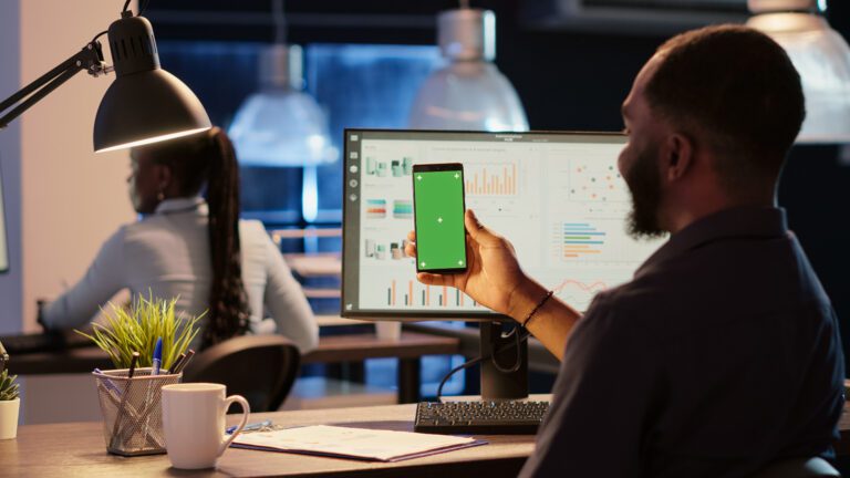 A man at a desk holds a smartphone with a green screen, navigating mobile apps, while his computer displays software development graphs. In the office setting, another person is working diligently in the background.