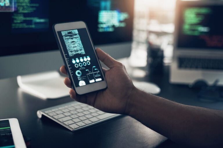 A person holding a smartphone displays code related to app development, standing before computer screens filled with intricate lines of programming.