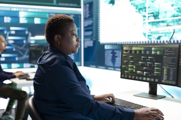 In a high-tech control room, a person works diligently at a computer with multiple screens displaying complex data and graphs, focused on threat detection and response.