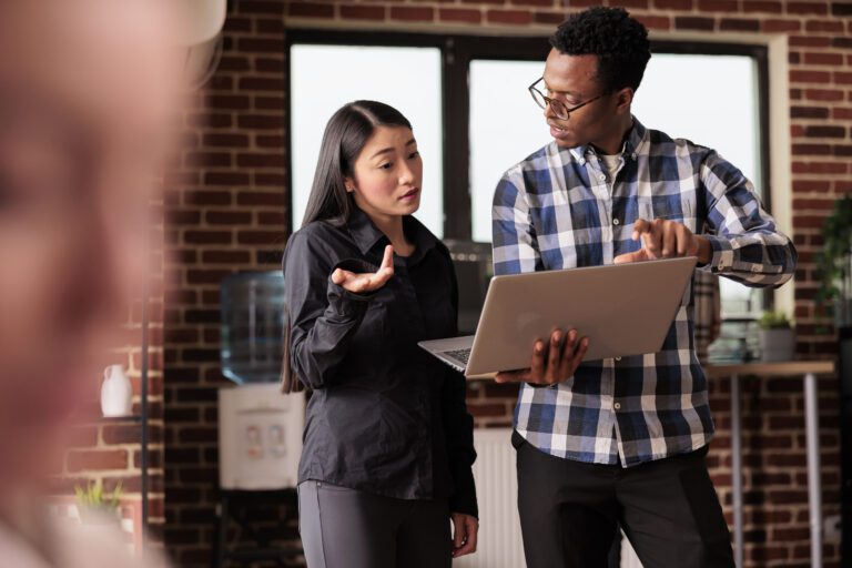 Two people engage in a discussion about risk management while examining data on a laptop in their office, framed by rustic brick walls.