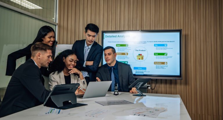 A group of professionals in a meeting room discuss app development strategies while looking at a laptop, with a presentation displayed on a screen behind them.