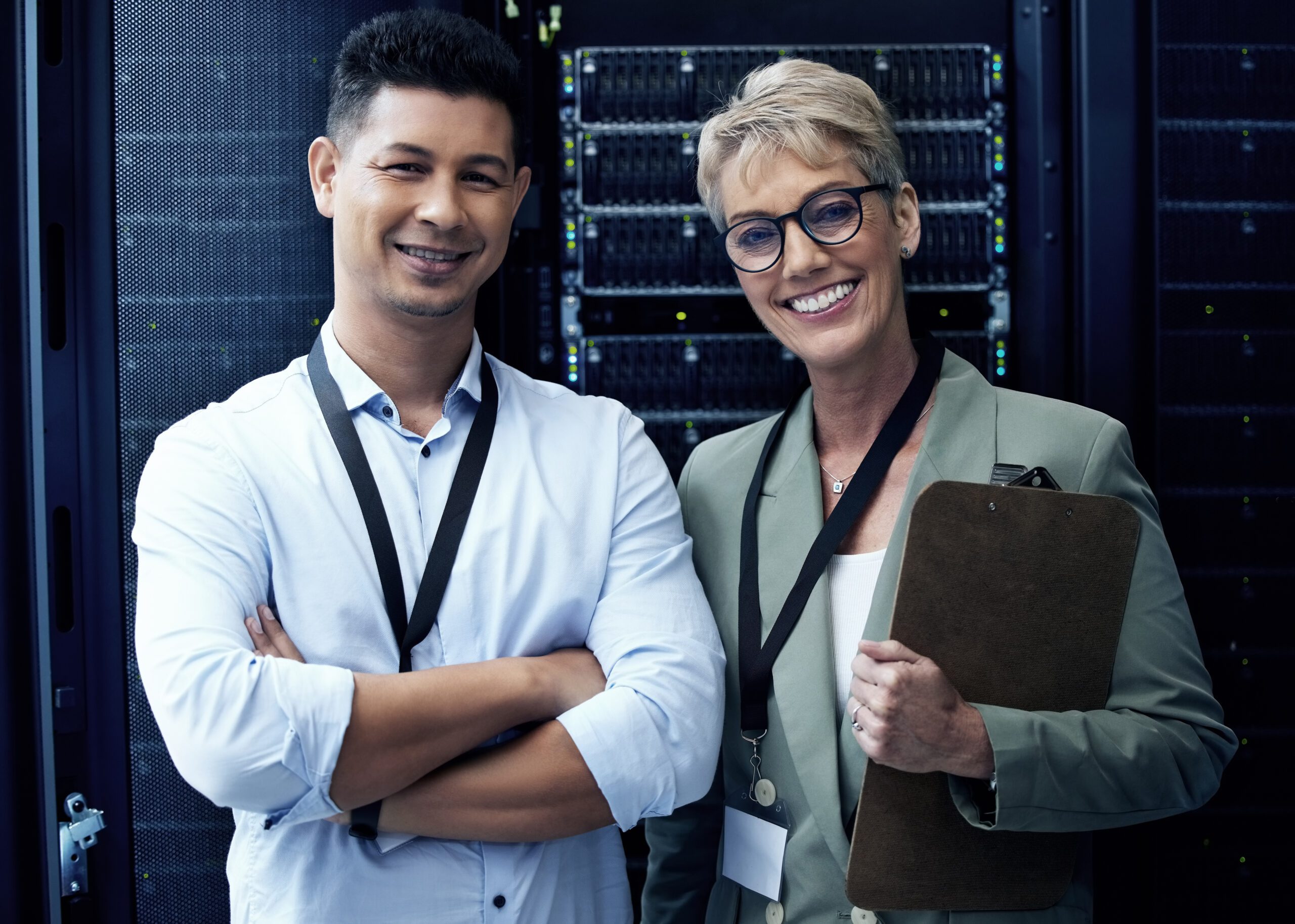 In a bustling server room, two people stand smiling, their ID badges catching the light. One holds a clipboard, a testament to their role in providing top-notch Managed IT Services.