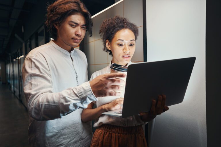 Two people stand in a hallway, focused on a laptop screen. One holds a coffee cup, discussing network security as they ensure secure connectivity while managing data protection.