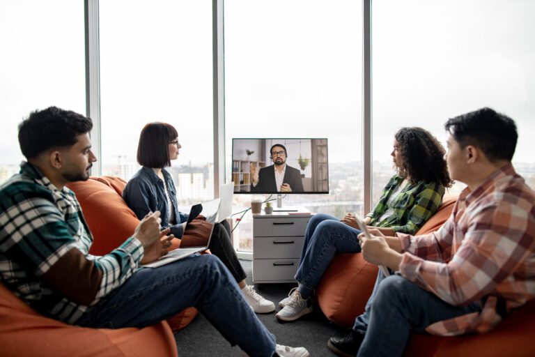 Four people seated in bean bags are watching a person on a video call displayed on a monitor in a modern office with large windows, seamlessly connected through their virtual desktop setups.