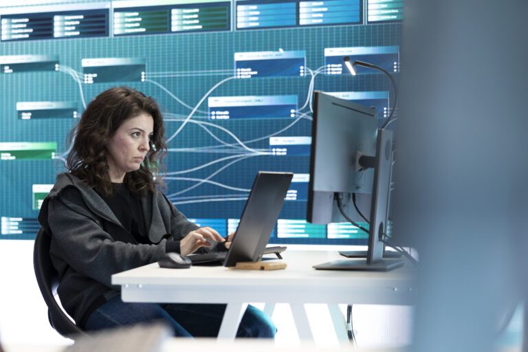 A woman working on a computer with data visualizations related to threat detection displayed on a screen in the background.