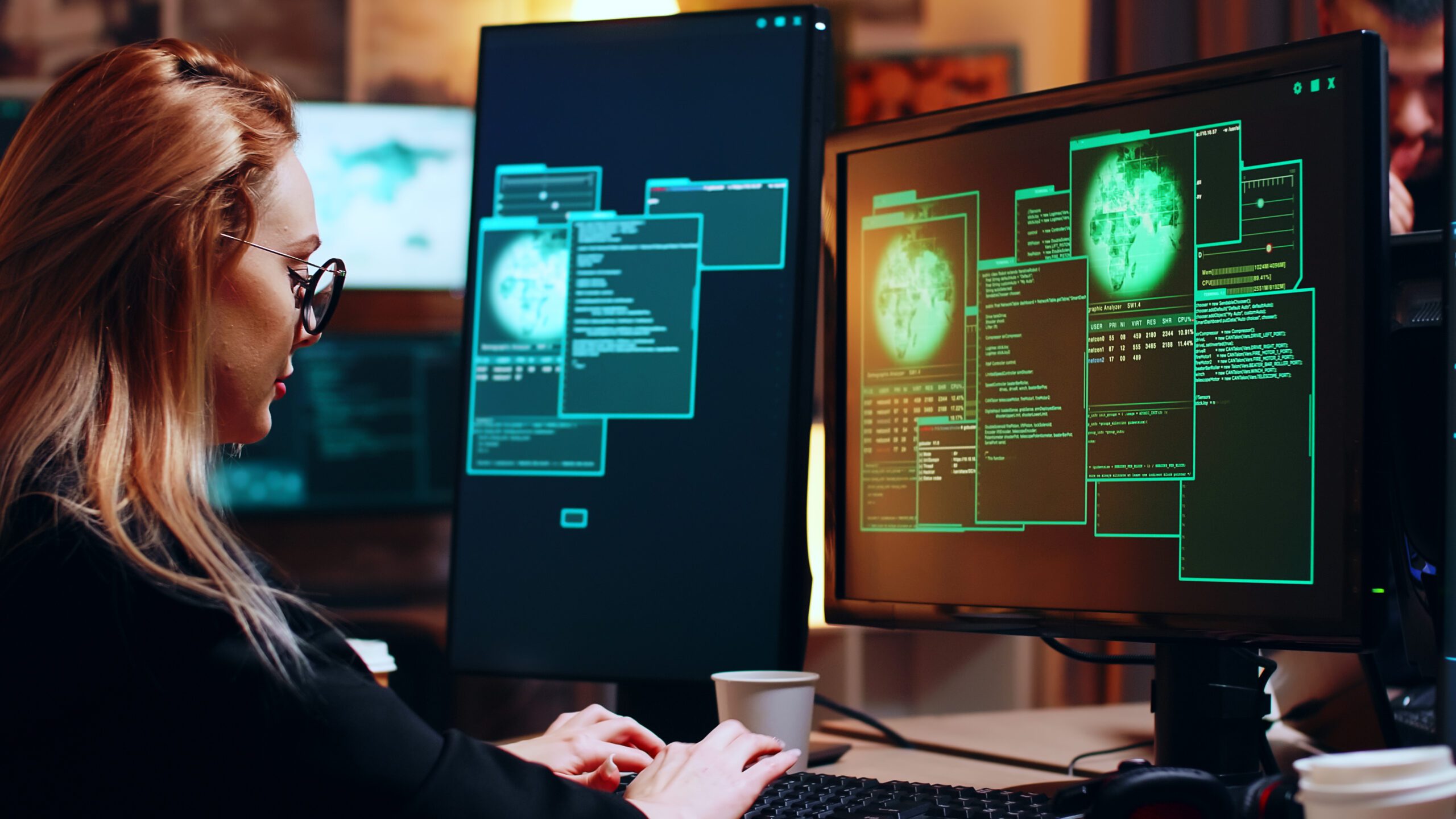 A person is working diligently at a desk, surrounded by multiple computer monitors displaying digital code and graphics, focused on threat detection and response.