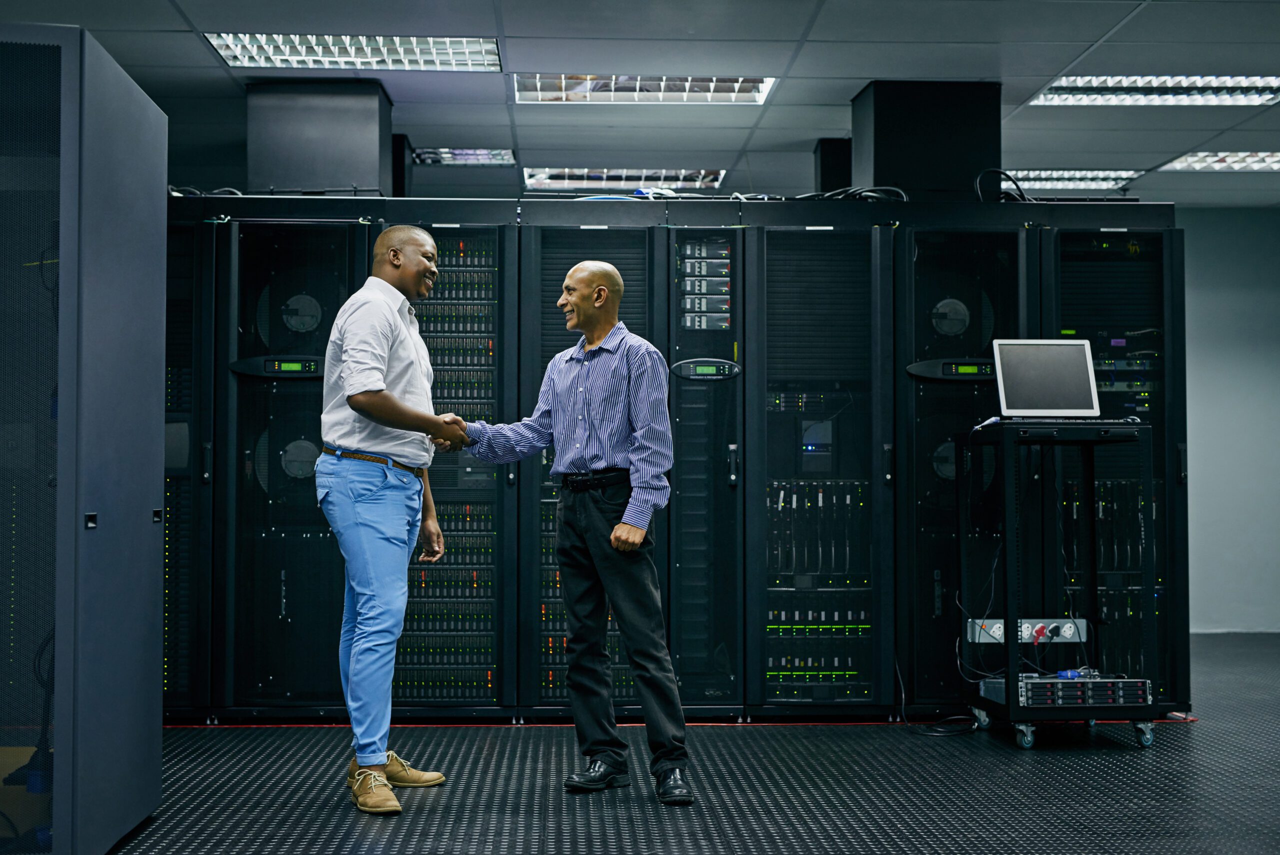 Two men shaking hands in a server room, surrounded by computer racks and a laptop on a stand, symbolizing the seamless integration of Hybrid Cloud solutions.