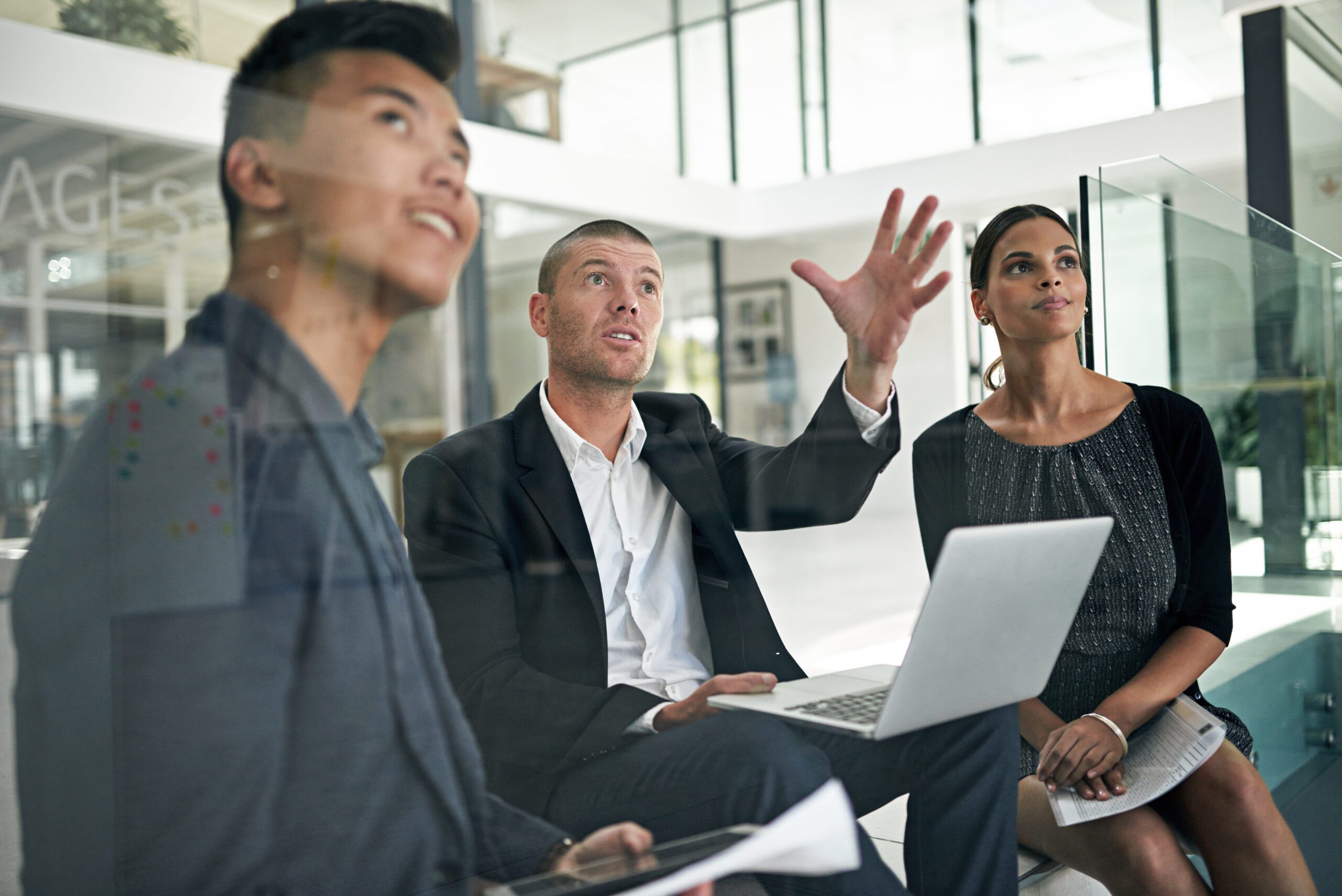 Three professionals sit in a modern office. One man, gesturing upward while holding a laptop, discusses strategies on how to transform your business. The other two individuals observe attentively, eager to embrace the potential for business transformation.