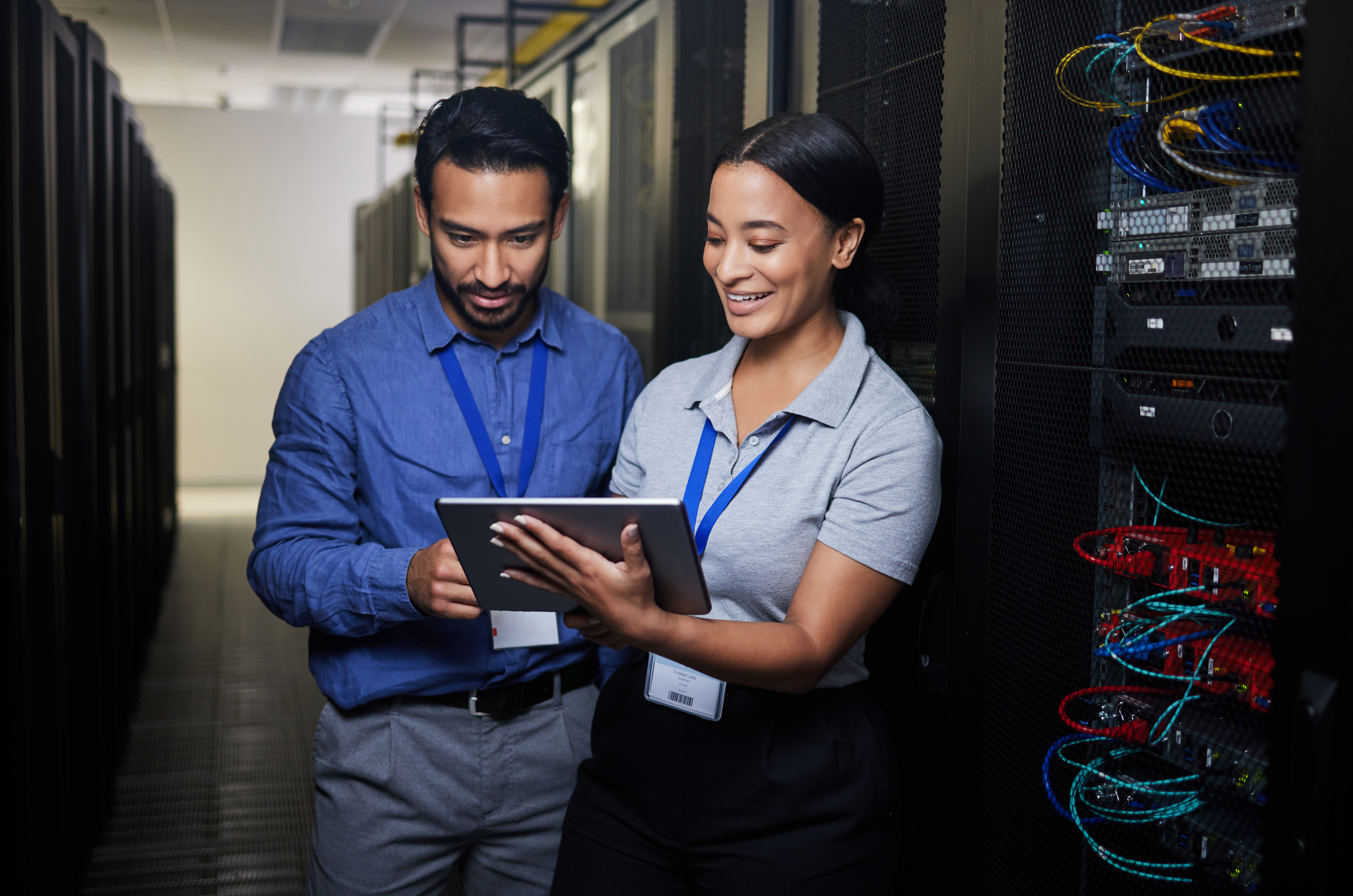 Two people in a server room, wearing badges and smiling, explore the potential of hybrid cloud solutions on a tablet.