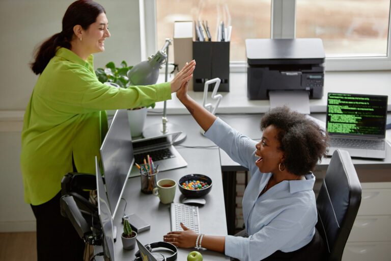 Two women high-fiving in an office near computers and other desk items, including a bowl of candies, an apple, and various collaboration tools.