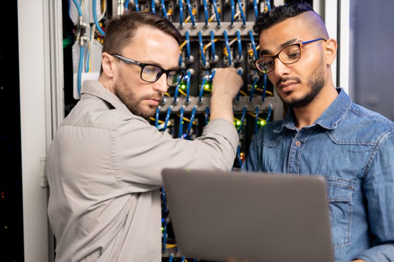 Serious pensive young multi-ethnic server specialists in glasses standing in database room and testing network system using laptop