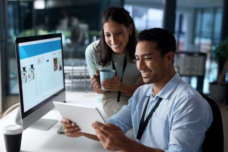 Two colleagues in an office setting review cloud solutions on a tablet together. One holds a mug, while the other sits at a desk with a computer monitor, discussing potential management solutions for their team.