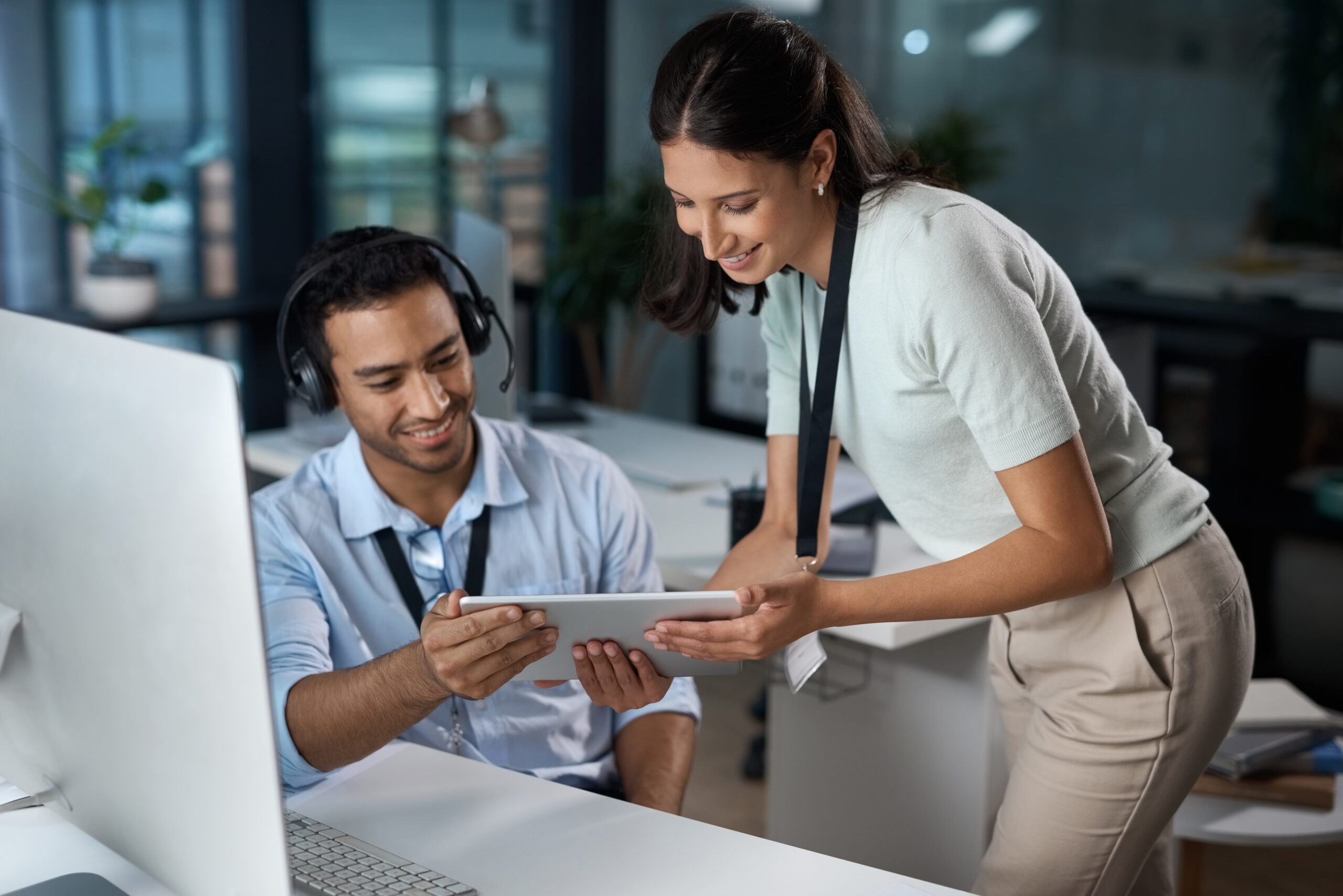 A man with a headset sits at a desk using a tablet, exploring IT management solutions, while a woman stands next to him, smiling in an office environment.