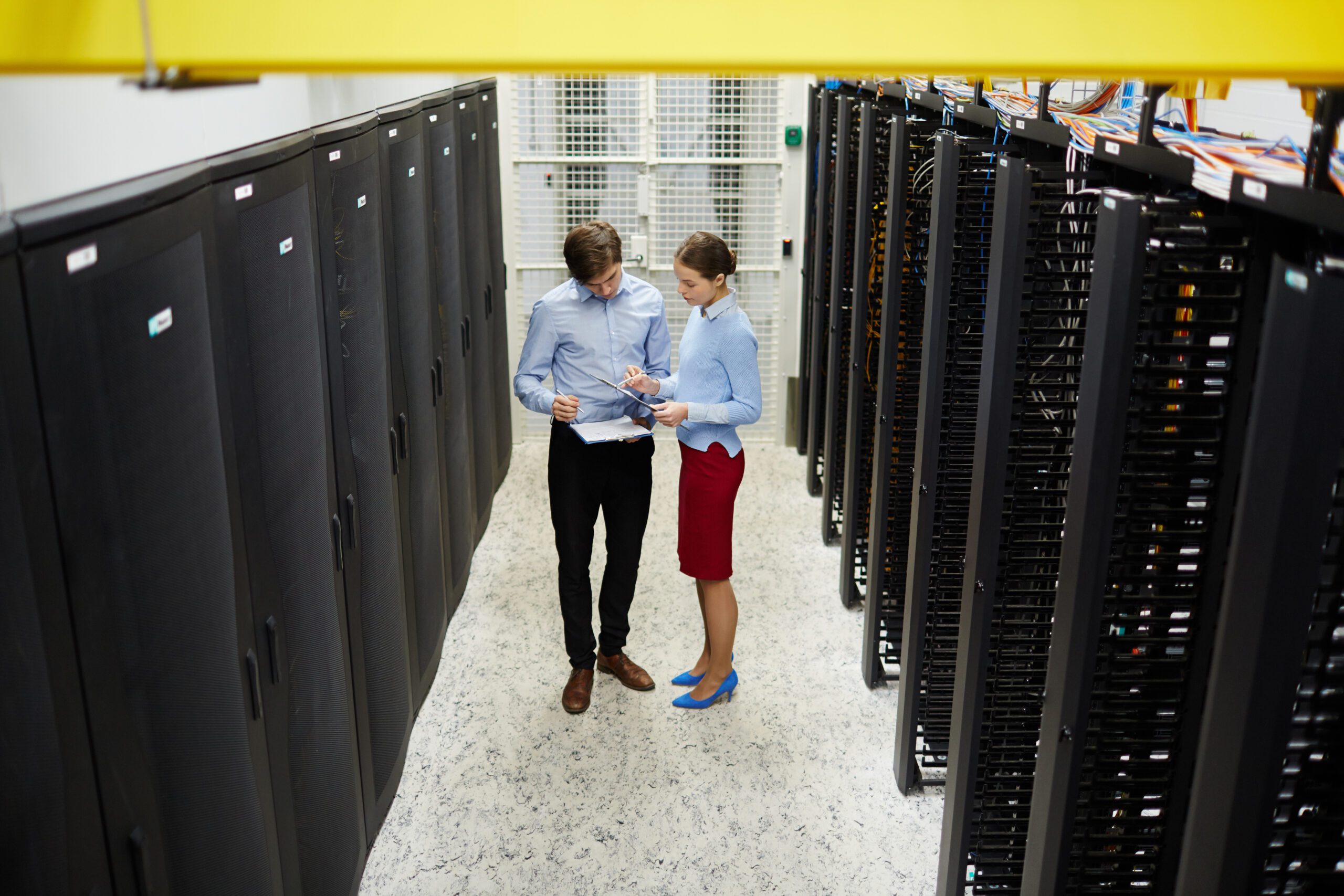 Two people stand in a server room, examining documents. Surrounded by tall black server racks on either side, they discuss ways to modernize the infrastructure, ensuring it meets future technological demands.