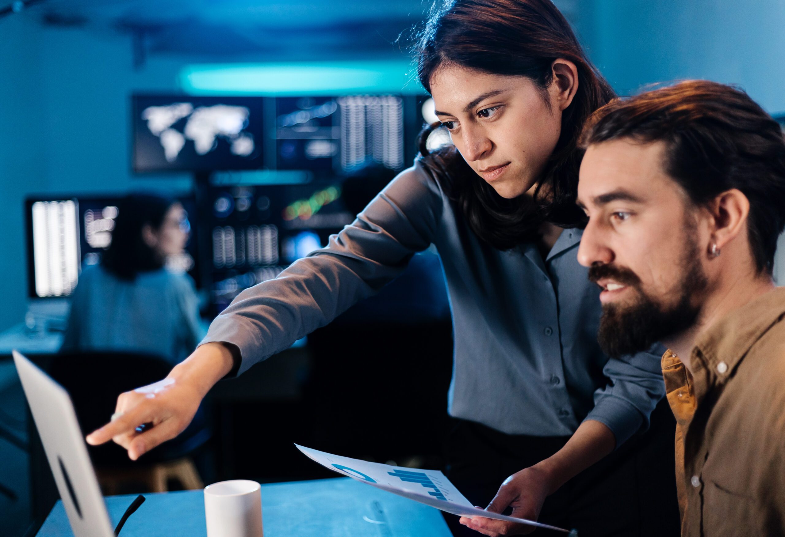 Two people in an office setting focus intently on a computer screen. One person points at the screen while holding a document with graphs, discussing cybersecurity strategies. Monitors displaying data and threat detection metrics are visible in the background, enhancing their response planning.