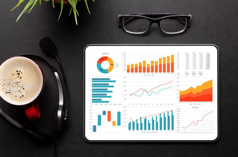 The tablet intelligently displays various colorful graphs and charts on a desk, surrounded by glasses, a cup of coffee, a headset, and a plant—creating an ideal setup for diving into deep analytics.