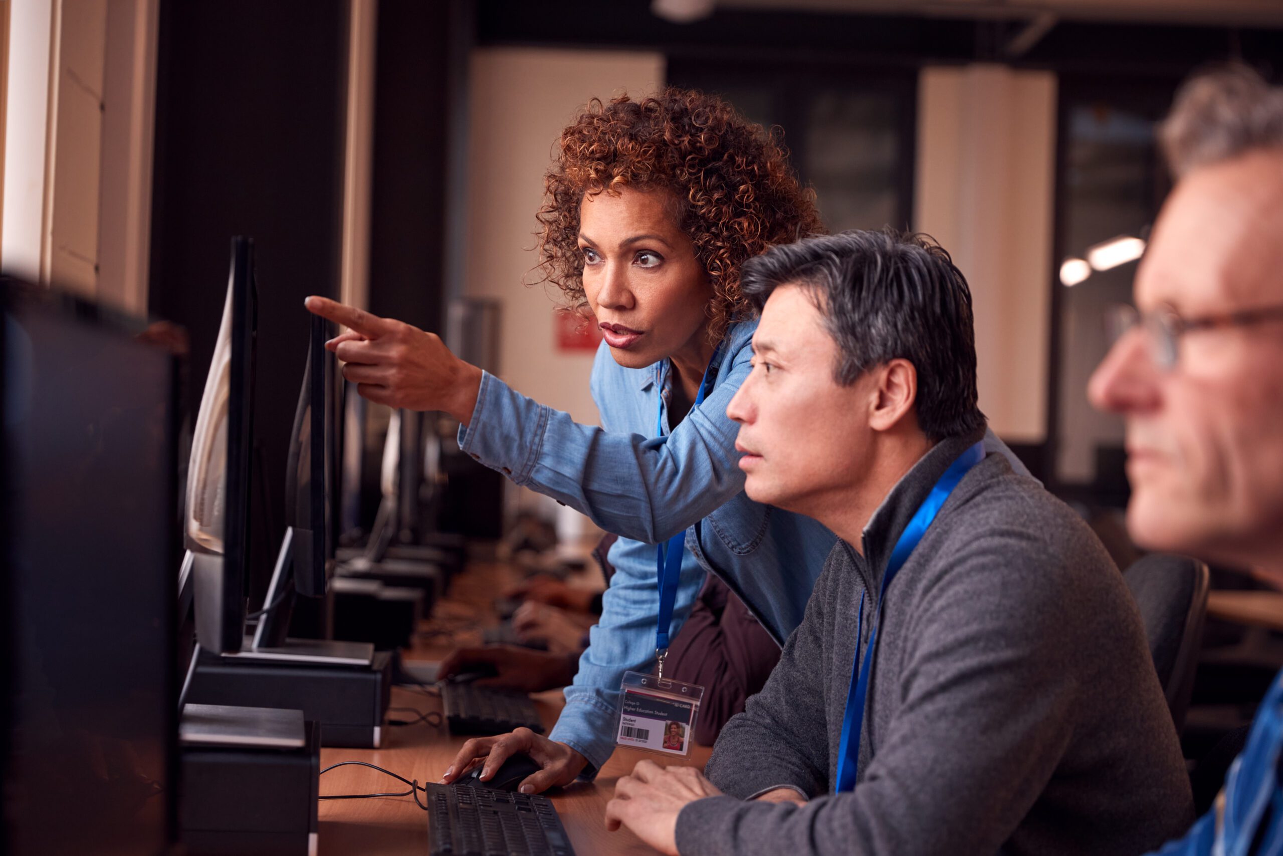 A woman points at a virtual desktop on the computer screen while assisting two men in an office setting.