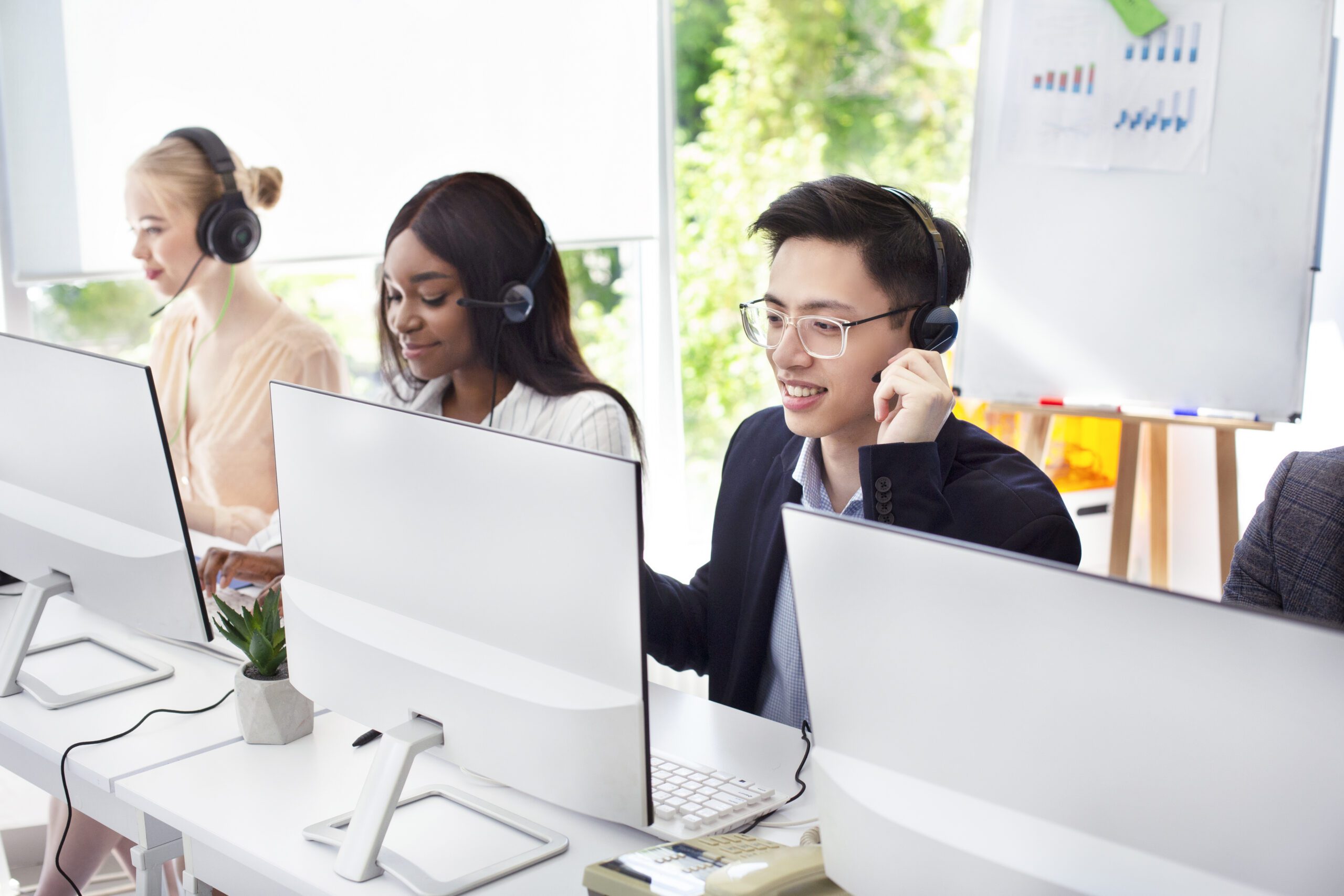 Four people are sitting at desks, wearing headsets, and working on computers in a bright office setting. A whiteboard with charts in the background showcases their focus on IT solutions and strategies for effective management solutions.