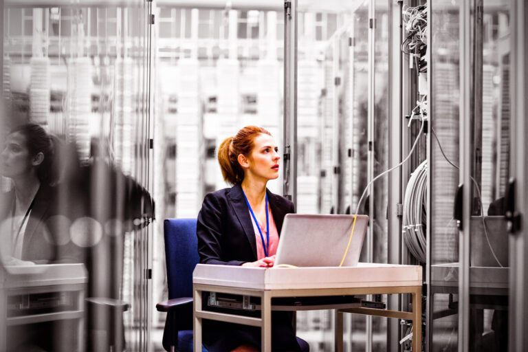 A person seated at a desk in a server room, working on a laptop amidst the visible cables. The glass walls reflect the complexity of managing a hybrid cloud environment.