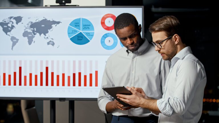 Two men engaged in an intelligent discussion about analytics, examining data on a tablet in front of a presentation screen showcasing a world map, pie charts, and a bar graph.