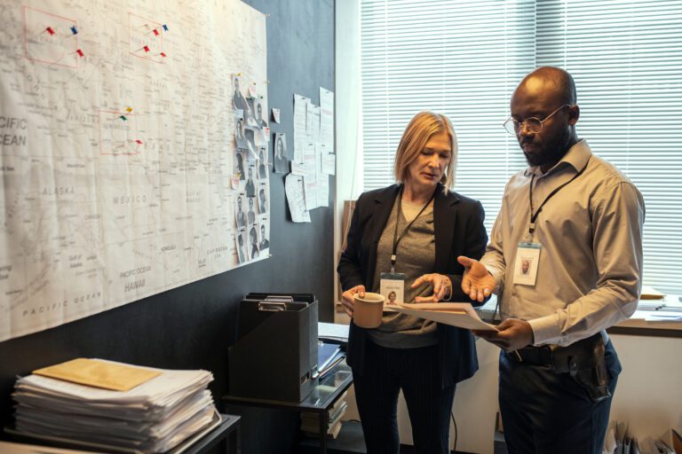 Two confident agents of FBI in formalwear discussing criminal profiles of suspects while standing in front of board with map