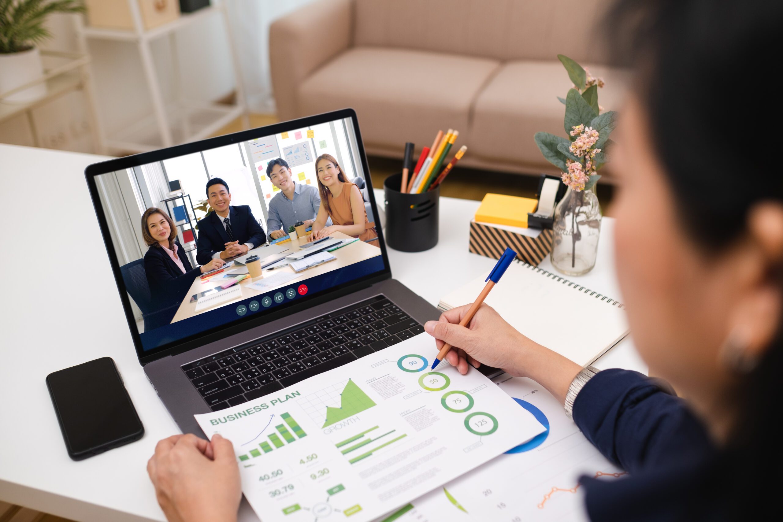 Person on laptop video call with four colleagues, showcasing a report with charts. Office supplies and notes are scattered on the desk, enhancing teamwork and improving collaboration among participants.