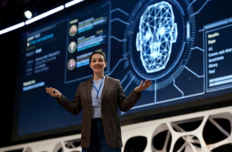 A person is giving a presentation on a stage, seamlessly blending insights as a large screen displays an AI-generated digital face and data, accelerating the audience's understanding of cutting-edge technology.