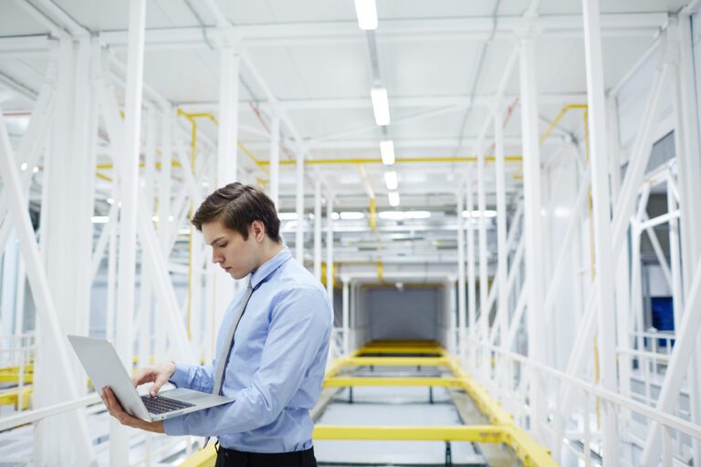 A person in a blue shirt efficiently manages IT solutions on their laptop while standing in a large industrial facility with white beams and yellow accents.