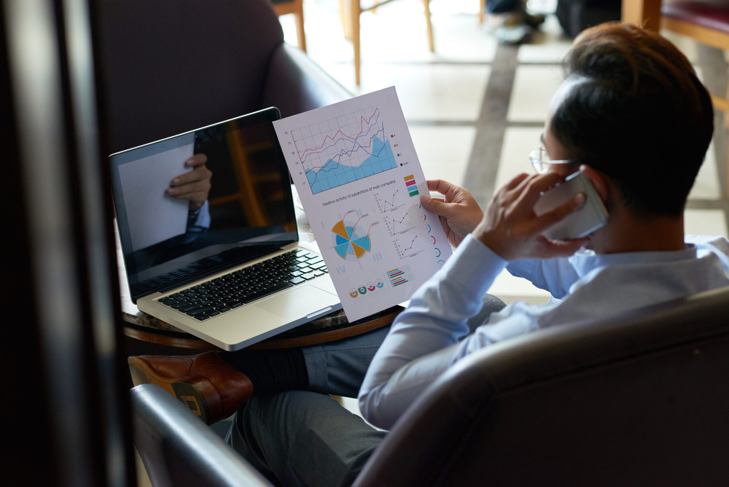 A person in a blue shirt is deeply engaged in intelligent analytics, speaking on the phone while examining charts and graphs, with a laptop open in front of them.