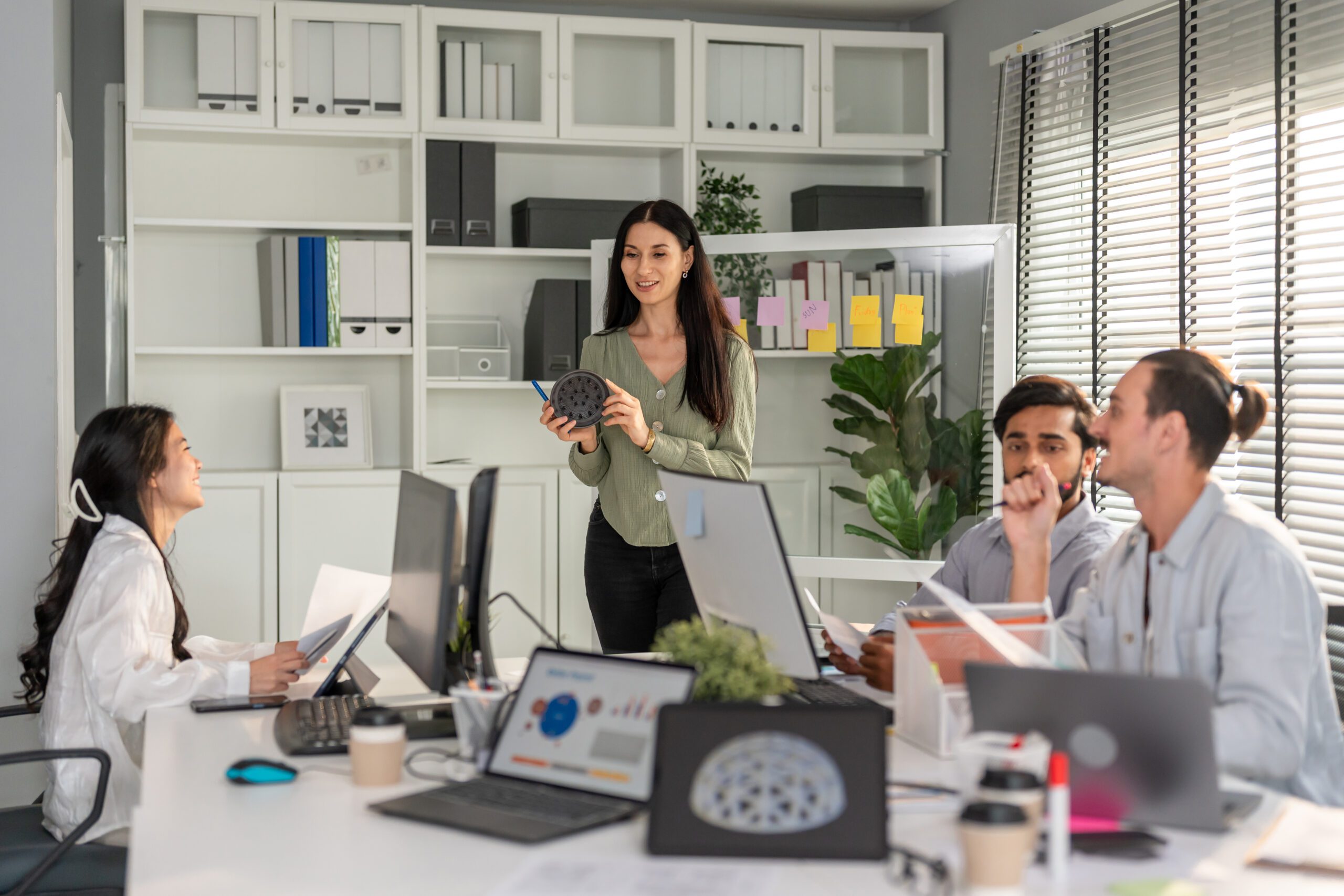 In an office setting, collaboration is evident as three people work intently at a desk laden with laptops and documents, while another stands holding a device. The room, equipped with tools such as shelves and window blinds, fosters a productive environment.