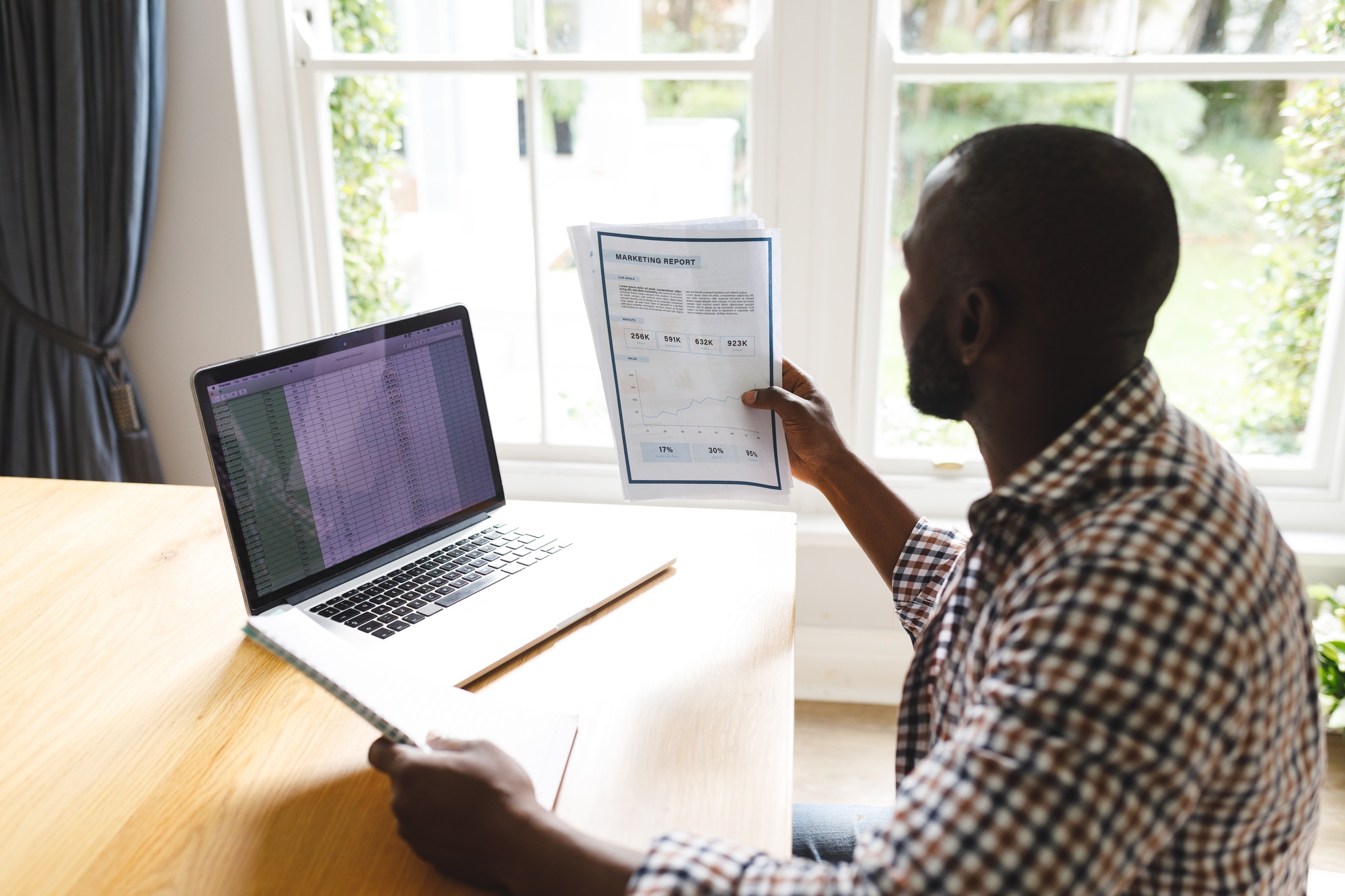 African american man sitting at table with paperwork in dining room, working remotely using laptop