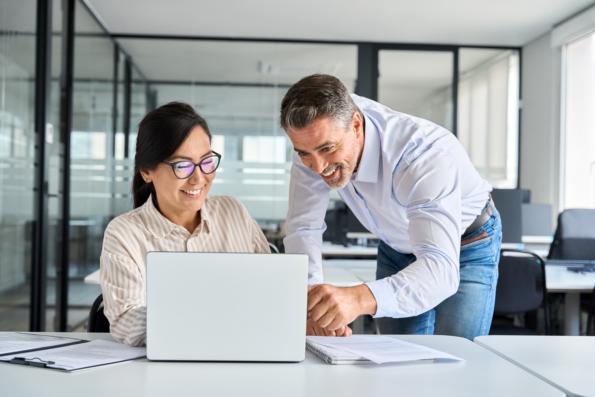 Mature boss manager helping Asian employee working on laptop in office.